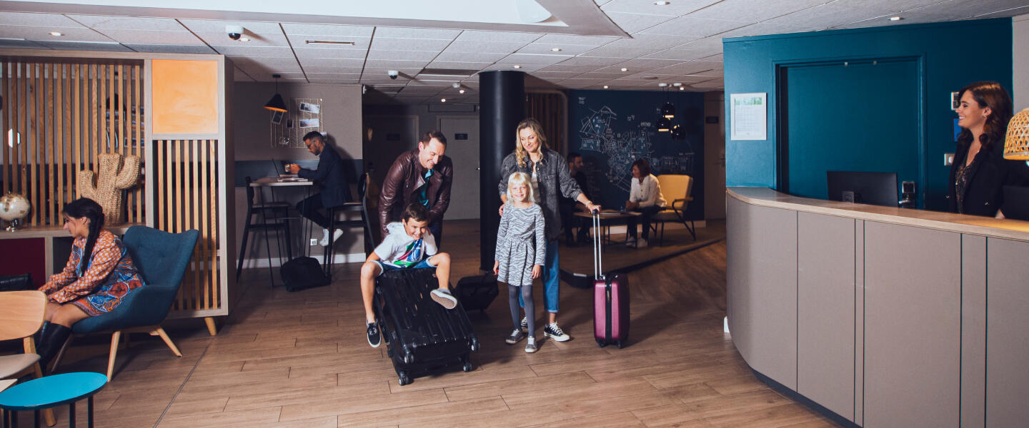 Famille avec enfants et bagages arrivant à la réception d'un appart'hôtel moderne et accueillant, avec des invités travaillant et se relaxant dans le lobby, illustrant la garantie du meilleur prix